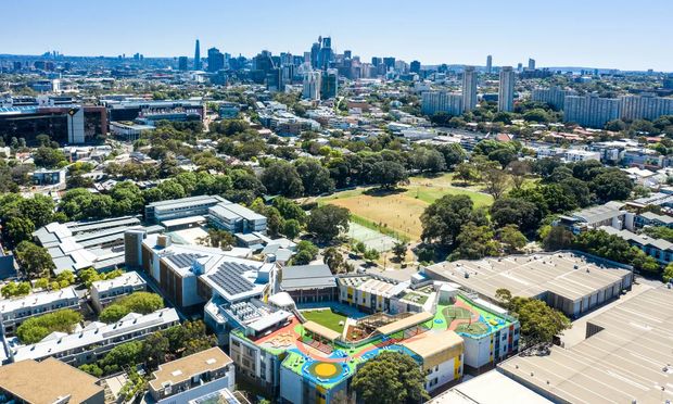 A high-angle aerial view of a dense urban area, featuring a large school or institutional campus in the foreground with colourful rooftop play areas, surrounding sports fields and mature trees, and a prominent city skyline in the distance. The scene is captured in bright daylight with clear blue skies.