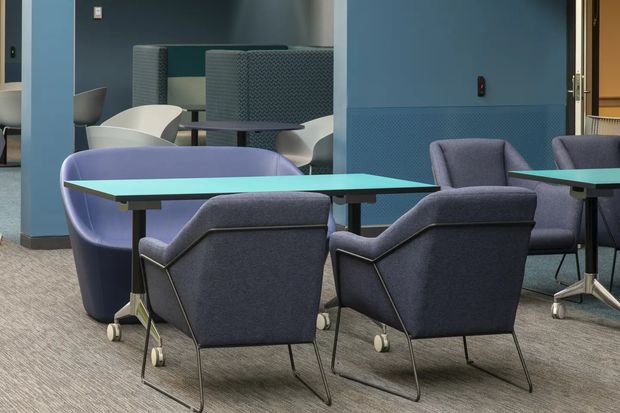 A contemporary commercial lounge or breakout area with blue upholstered armchairs arranged around turquoise rectangular tables on casters. The space features blue wall panels, acoustic booth seating in the background, grey carpet flooring, and a partially visible adjoining seating area. The furnishings and finishes create a refined, cool-toned workplace or hospitality setting.