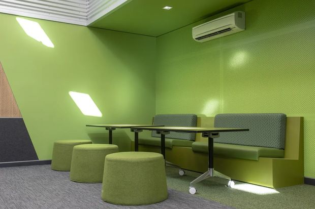 A compact, brightly coloured commercial-style lounge or breakout space with vivid green walls and ceiling, perforated acoustic wall panels, built-in upholstered bench seating, a long dark table, and several rounded green ottomans. A wall-mounted split-system air conditioner and recessed ceiling lighting are visible. The room has grey carpet flooring and a bold, playful, contemporary fit-out.
