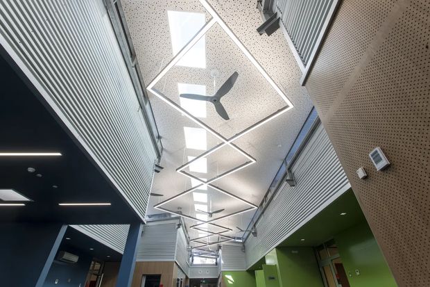 Interior view of a contemporary multi-level commercial or institutional atrium with a tall ceiling, exposed upper walkways or room fronts, extensive acoustic perforated wall and ceiling panels, linear ribbed metal screens, skylights, and geometric suspended LED light fixtures. A large ceiling fan and bright green wall accents add visual interest to the otherwise neutral grey, white, and black palette.