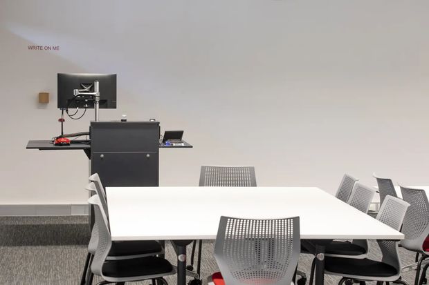 A clean, modern commercial meeting or collaborative workspace with a large white rectangular table surrounded by ergonomic mesh chairs. At the front is a wall-mounted display and presentation station with a dark equipment cabinet, monitor, keyboard shelf, and visible cables. The room has plain light walls, grey carpet, and a small “WRITE ON ME” wall label.