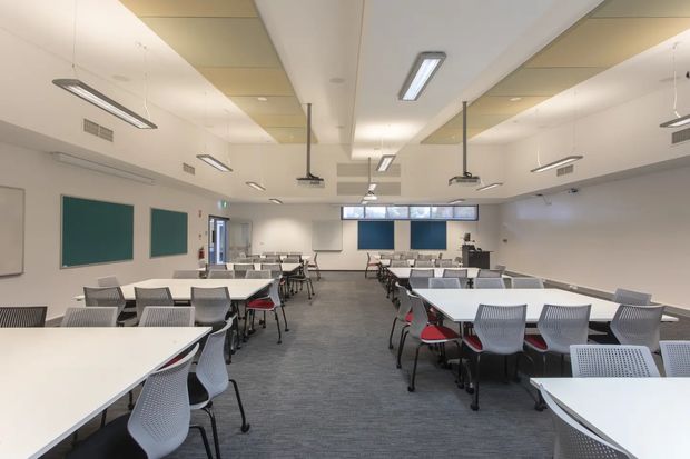 A large contemporary classroom or training room with rows of white tables and grey perforated chairs, several red accent chairs, teal acoustic wall panels, suspended linear light fixtures, ceiling-mounted projectors, and grey carpet flooring. The space is bright, orderly, and designed for flexible group learning or presentations.