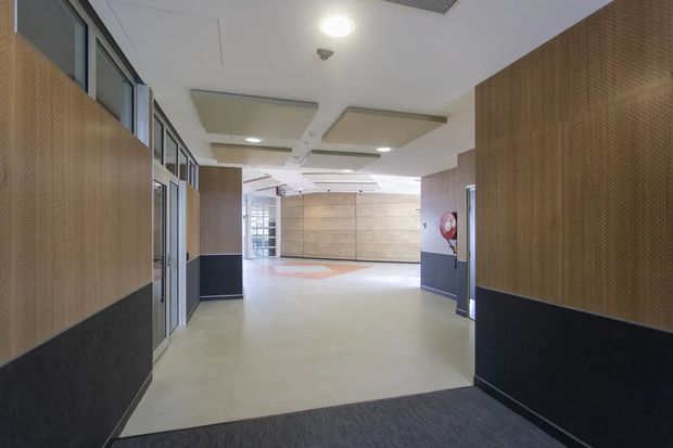 A modern institutional or commercial interior hallway opening into a spacious lobby. The corridor features perforated timber wall panels with dark lower sections, glazed office or room fronts along the left side, pale polished flooring, acoustic ceiling panels, recessed lighting, and a large timber-clad wall in the background. A fire hose reel is visible on the right wall.
