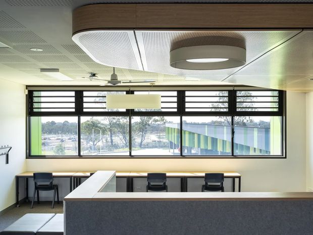 Bright contemporary commercial interior, likely an open-plan office or educational workspace, viewed from behind a large dark reception or counter-height partition. A long run of desks with task chairs lines the glazed wall, with additional seating at a perpendicular workbench. The room features expansive horizontal black-framed windows, acoustic ceiling panels, integrated linear lighting, and a sculptural curved timber-and-white ceiling bulkhead. Trees and a colourful neighbouring facade are visible outside.