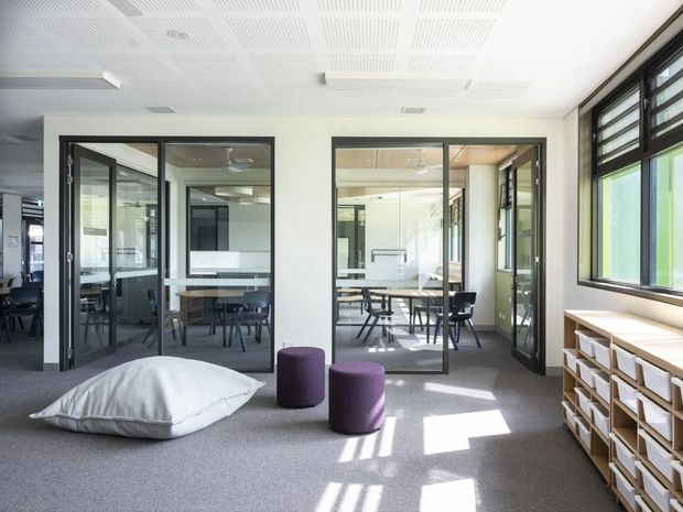 Bright contemporary commercial interior with a flexible breakout lounge and meeting-room spaces. The foreground has a grey carpeted floor, oversized white floor cushion, and two purple cylindrical ottomans. Black-framed glazed partitions enclose meeting rooms with tables and chairs, while large black-framed windows provide strong daylight. Open shelving with storage boxes lines the right wall, and the ceiling has acoustic perforated panels with integrated linear lighting.