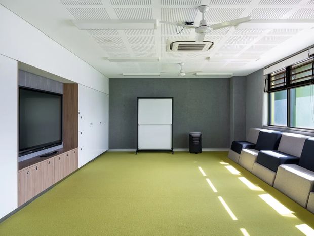 Bright, contemporary multipurpose room with a lime-green carpet, dark grey textured feature wall, integrated timber-look media cabinetry, rolling whiteboard, and a row of upholstered lounge chairs beneath large windows with timber louvres. The ceiling has acoustic perforated panels, recessed lighting, and ceiling fans. The space appears suitable for meetings, presentations, or informal group gatherings.