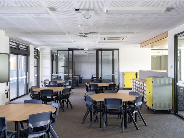 A bright, open-plan commercial office or educational workspace with multiple round timber meeting tables and black chairs. The room has extensive black-framed glazed partitions and doors, a carpeted floor, white acoustic perforated ceiling panels with linear fluorescent lighting, ceiling fans and air-conditioning. Yellow and grey filing or storage units line the right side, with additional workspaces visible through the glazed partition.