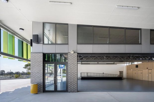 A modern commercial or community facility with a covered open entrance and large internal hall. The facade combines dark grey brickwork, black-framed glazed doors and clerestory windows, and grey metal wall panels. Inside, a broad polished grey floor, exposed roof trusses, mezzanine-style railings, and pale timber acoustic wall panelling are visible. Bright green and blue vertical exterior panels add colour along the left side.