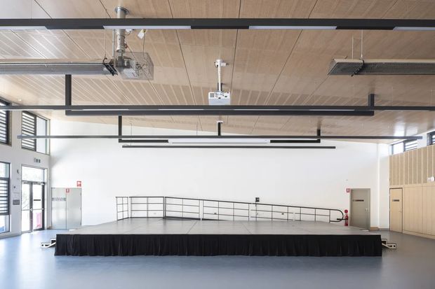 A bright, spacious multi-purpose hall or commercial event space with a low black stage at the front, open circulation area, high exposed services, suspended linear lighting, pale timber ceiling panels, white walls, grey flooring, high-level windows, and light timber wall panelling. The space appears functional and contemporary, with visible heating or ventilation equipment and accessible entry doors.