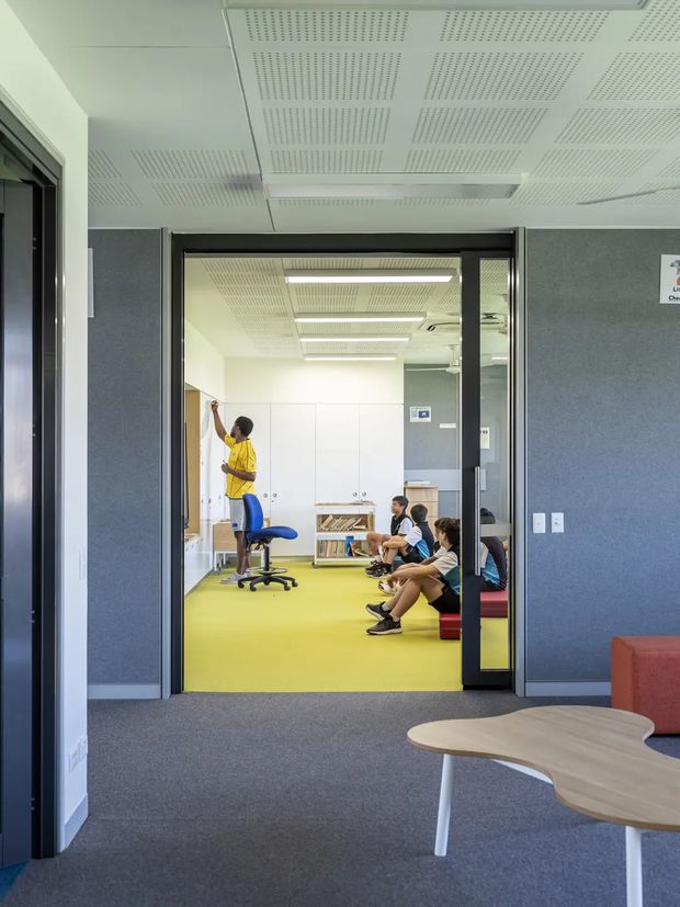 View through a glazed doorway into a bright institutional or educational room. The foreground is a carpeted corridor or waiting area with dark acoustic wall panels, a light timber-toned irregular table, and red upholstered seating. Beyond the black-framed glass doors is a colourful activity or classroom space with a vivid yellow-green floor, white built-in cabinetry, bookshelves, a blue office chair, and seated children. The ceiling throughout has white perforated acoustic panels and linear fluorescent lighting.
