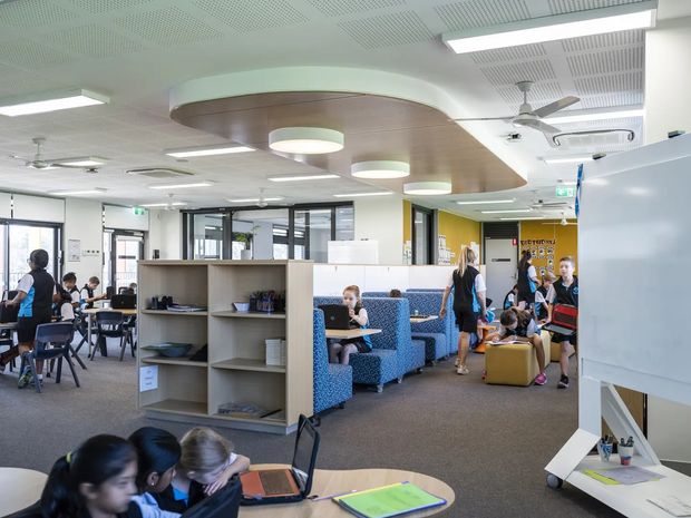 Bright, contemporary open-plan educational or collaborative workspace with students and adults seated at tables and in blue upholstered booth seating. The room features carpeted flooring, extensive internal glazing, open shelving, whiteboards, suspended linear lighting, ceiling fans, and a curved timber-look ceiling feature with integrated pendant lights. A yellow feature wall and colourful furnishings add visual interest.