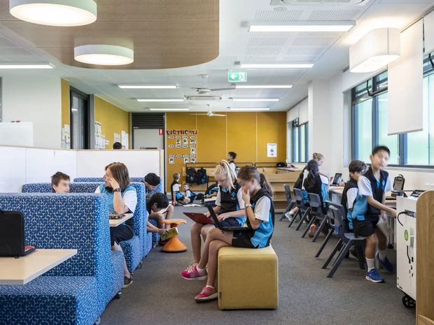 Bright, contemporary school or institutional learning commons with students seated at upholstered booth seating, tables, loose chairs, and ottomans. The open-plan room has large windows, a mustard-yellow accent wall, blue patterned acoustic booth upholstery, grey carpet, white acoustic ceiling panels, and suspended and recessed lighting. Workstations and educational display boards are visible throughout.