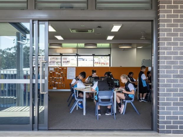View through a wide glazed entrance into a bright classroom or collaborative learning space. Students sit around tables and work independently or in small groups, with an orange pinboard wall, whiteboard, large windows, fluorescent ceiling lights, and dark carpet. The exterior opening is framed by dark brick and aluminium-framed glazing.