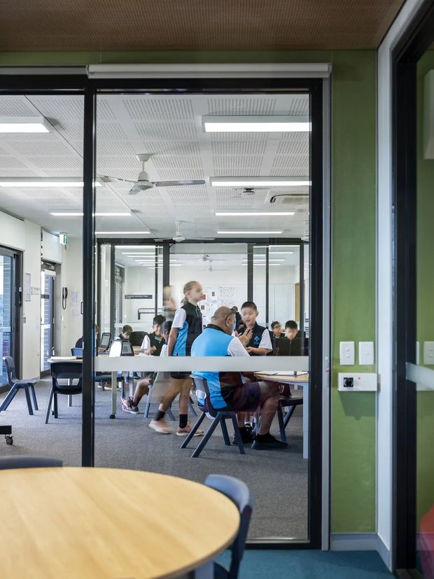 Interior view of a contemporary educational or commercial meeting space, photographed through black-framed glazed partitions. Students and staff are gathered around tables in an open collaborative room. The space has green painted walls, carpet flooring, acoustic perforated ceiling panels, linear fluorescent lighting, ceiling fans, air-conditioning, and durable institutional furniture.