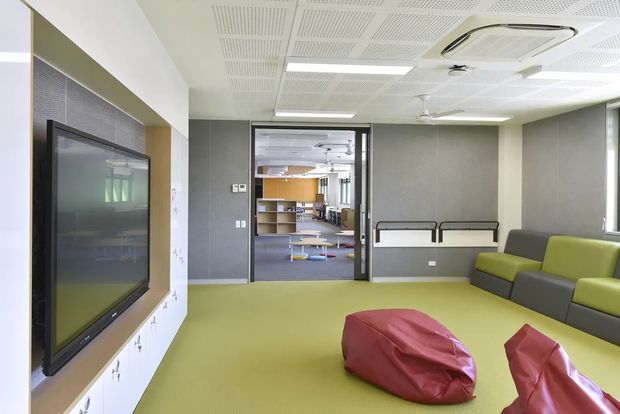 A bright contemporary commercial interior, likely a flexible lounge or breakout area, with lime-green flooring, grey walls, modular grey-and-green seating, red beanbag chairs, a wall-mounted display, and an open doorway leading to an adjacent workspace or collaborative area. The room has a suspended acoustic ceiling with integrated fluorescent lighting and ceiling-mounted air conditioning.