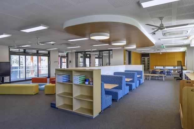 A bright contemporary commercial interior, likely a school or institutional communal dining and breakout area. The open-plan room features blue upholstered booth seating with timber tables, yellow and orange lounge seating, built-in shelving, a yellow accent wall, extensive glazing, carpeted flooring, acoustic perforated ceiling panels, suspended linear lighting, ceiling fans, and exposed air-conditioning units. A rounded timber-lined ceiling feature with integrated pendant lights forms a central architectural focal point.