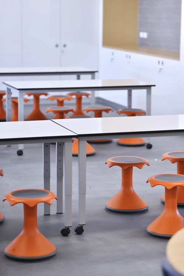 A bright, modern classroom or flexible learning space with rows of white rectangular tables on castors and distinctive orange, pedestal-style stools. The room has a smooth grey floor, white built-in cabinetry, and a large timber-toned wall panel, creating a clean, functional educational environment.