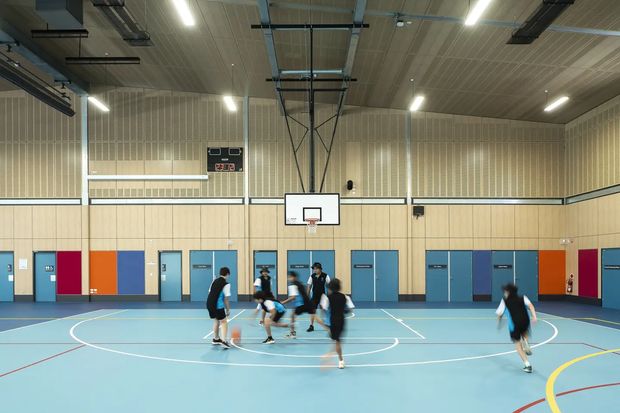Bright indoor basketball court in a large multipurpose sports hall, with a pale blue sports floor marked by basketball and other court lines, wall-mounted hoops, a scoreboard, timber-toned acoustic wall panelling, high exposed services, and several players. Blue, orange, red, and grey doors and panels line the perimeter walls.