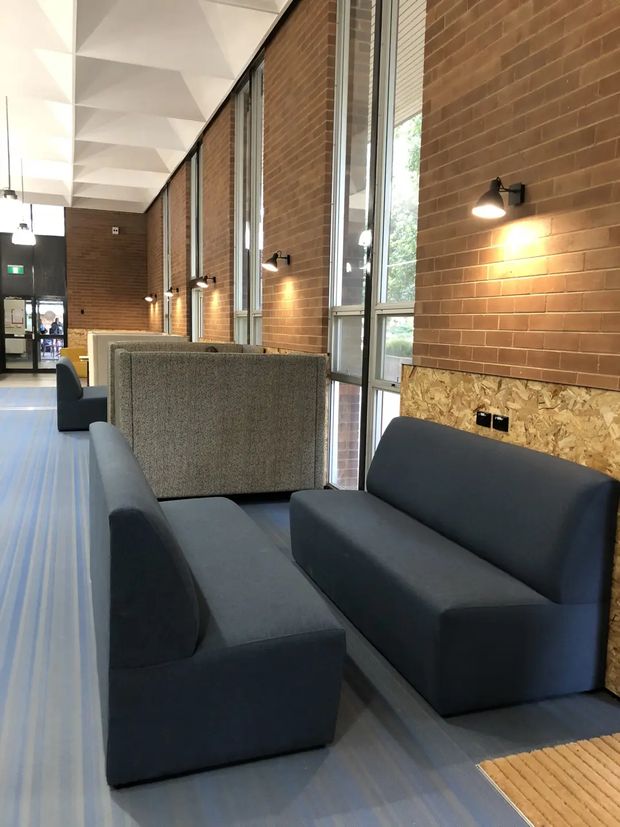 A contemporary commercial interior lobby or lounge with dark upholstered sofas arranged along a long exposed brick wall. Tall aluminium-framed windows provide natural light, while black wall-mounted sconces illuminate the seating area. The ceiling features a sculptural white geometric treatment, and the floor is covered with striped carpet. OSB-style timber paneling appears as a lower wall finish behind the seating.