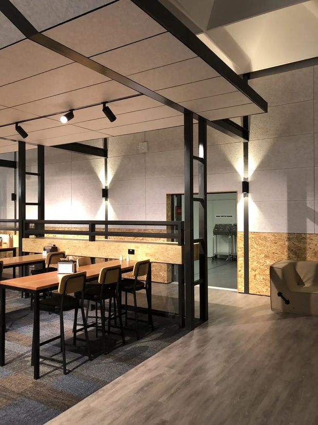 A contemporary commercial interior resembling a café, restaurant, or hospitality dining space. The room features communal and individual timber tables with black metal frames, upholstered dining chairs, a high counter-height table, black steel-framed partitions and beams, pale concrete-look wall panels, plywood wall wainscoting, exposed structural detailing, track lighting, and warm wall sconces. A separate service or back-of-house area is visible through an opening, while a lounge chair occupies the right foreground.