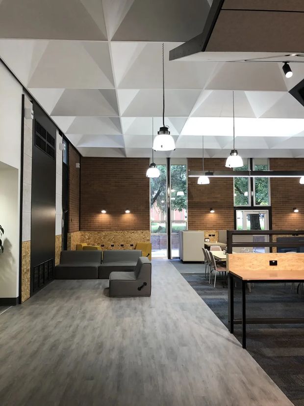 A contemporary commercial interior, likely a flexible office or collaborative workspace, with a dramatic faceted white geometric ceiling, exposed dark structural elements, suspended globe pendant lights, dark brick walls, expansive windows, and an open-plan arrangement of sofas, desks, chairs, and partitioned work areas. The space uses a muted grey wood-look floor, warm timber-toned work surfaces, and dark metal furnishings.