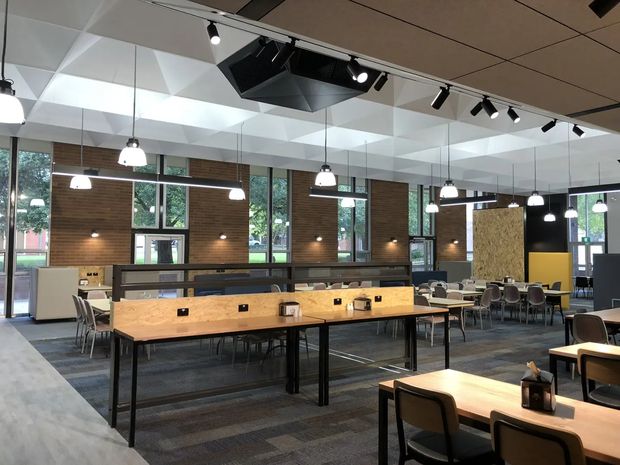 A spacious contemporary commercial dining and communal workspace with long timber tables, black metal frames, pendant lighting, and rows of upholstered chairs. The room features exposed acoustic ceiling panels, track lighting, extensive internal glazing, and a warm timber-panelled wall with large windows overlooking greenery. A service or kitchen counter is visible toward the rear.