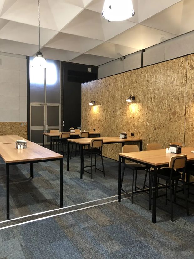 A contemporary commercial dining or café interior with several light timber tables and black metal-framed chairs arranged across a carpeted floor. A prominent OSB wood-panel feature wall runs along the right side, illuminated by small wall-mounted lights. The room has dark painted walls, glass doors, a suspended geometric acoustic ceiling, and pendant lighting, creating an industrial yet warm atmosphere.