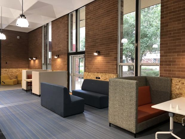 A contemporary commercial lounge or breakout area with high exposed red-brick walls, tall industrial-style windows, and a faceted white ceiling. The space contains upholstered booth seating and sofas in dark blue, grey, and rust tones, arranged on a striped carpet, with pendant and wall lighting providing a warm, functional atmosphere.