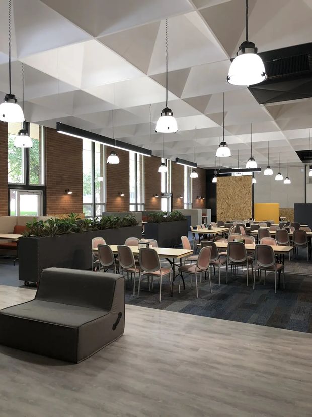 A large contemporary commercial dining and social space with rows of tables and chairs, black upholstered lounge seating, planter partitions, tall brick walls, extensive windows, and numerous suspended pendant lights. The ceiling features a dramatic geometric faceted white-panel design, while dark carpet tiles define the dining area and grey timber-look flooring occupies the foreground.