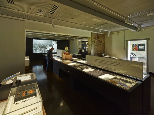 A dimly lit museum or exhibition gallery interior with a long central display island containing illuminated interpretive panels, archival documents, photographs, and artifacts. Dark polished flooring, muted olive-grey walls, a suspended acoustic ceiling with integrated services and track lighting, and wall-mounted exhibition graphics create a formal contemporary heritage-display environment. Visitors and display cases are visible throughout the space.