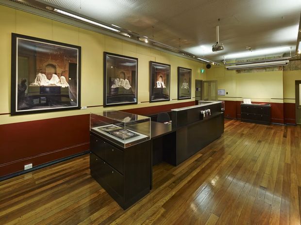 Interior view of a museum or gallery exhibition space with polished timber flooring, yellow-painted walls, dark red lower wall panelling, framed artwork, glass-topped display cases, and dark built-in counters. The room has a suspended ceiling with track lighting, ductwork, and a ceiling-mounted projector.