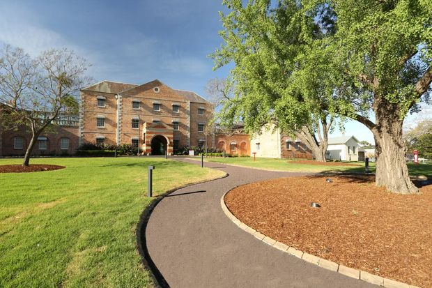A landscaped institutional or heritage-style brick building viewed from a curving paved walkway. The main structure is a multi-storey symmetrical brick building with a hipped roof, small multi-pane windows, and a prominent arched entrance. Lower brick wings and additional pale buildings extend around a broad lawn, mature trees, and mulched garden beds under a clear sky.
