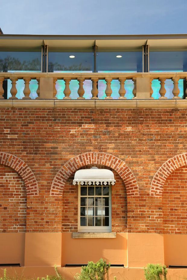 Close-up exterior view of a heritage-style brick facade. Red brickwork features decorative arched openings and a small multi-pane window with a projecting white curved awning. Above, a balustraded terrace with stone-colored balusters fronts a glazed upper level with a broad projecting roof or canopy. Warm terracotta-painted lower wall sections and a small amount of landscaping are visible at the base.