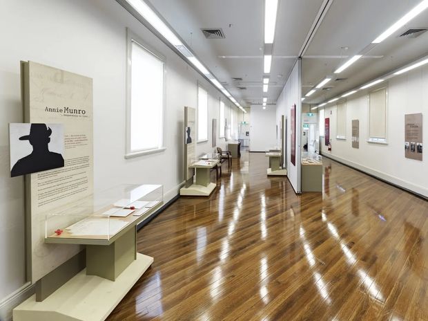 A bright, elongated museum or gallery exhibition interior with polished timber flooring, white walls, linear ceiling lighting, windows along one side, and freestanding display plinths holding framed educational panels and artefacts. The space has a clean, institutional contemporary fit-out and an open circulation layout.