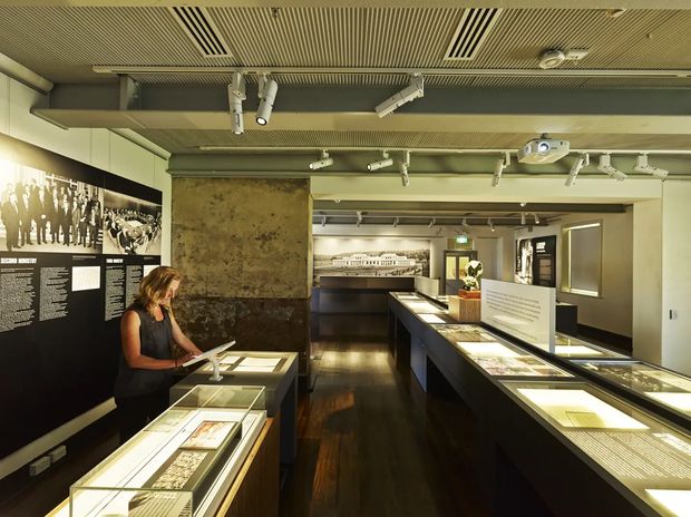 Interior view of a museum or heritage exhibition gallery with illuminated glass display cases, interpretive wall panels, archival documents, and photographs. The space has dark timber flooring, exposed structural beams and services, track lighting, and a linear slatted ceiling, creating a contemporary institutional exhibition fit-out.