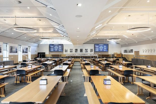 A bright, large contemporary commercial dining hall or cafeteria with many long timber tables and benches, black chairs, and numbered table signs. The room has a high ceiling with white acoustic panels, linear detailing, recessed downlights, and large circular pendant fixtures. A service counter and menu display screens line the far wall, with framed images and wall graphics adding visual interest. Grey carpeted aisles and neutral wall finishes create a clean, orderly institutional interior.