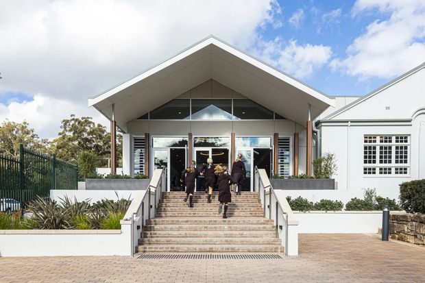 Front exterior view of a refined institutional or commercial building entrance. A contemporary gabled canopy with exposed timber-toned supports shelters broad glazed entry doors and a formal flight of tiled steps with metal handrails. The entrance is connected to a white classical-style wing with decorative window detailing, while landscaped planting, brick paving, and a dark metal fence frame the approach.