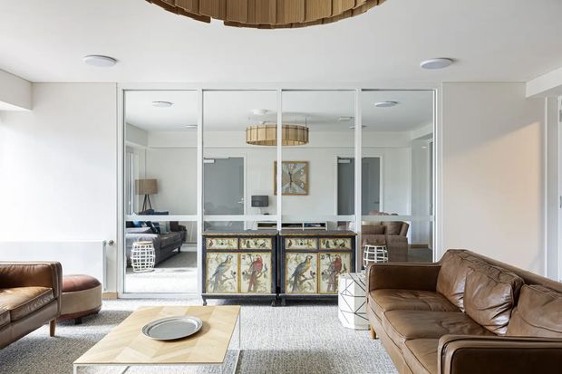 Bright contemporary living room with white walls and ceiling, large mirrored sliding or framed glass panels, a vintage-style painted cabinet, brown leather sofas and armchairs, a pale timber coffee table, and a woven pendant light. The mirrors reflect additional seating and the room's layered layout.