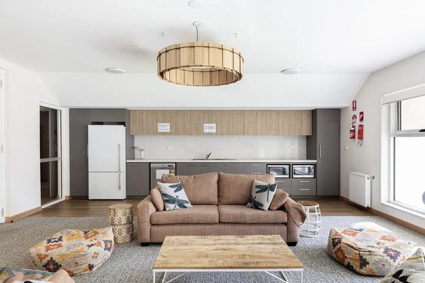 Bright open-plan living room with a compact integrated kitchen along the rear wall. The room features white walls and ceiling, dark grey lower cabinetry, light wood overhead cabinetry, a white refrigerator, timber-look flooring, a large woven pendant light, a neutral sofa, patterned cushions, a light timber coffee table, and colourful patterned floor cushions.