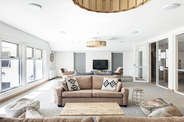 Bright contemporary living room with a large neutral upholstered sofa, patterned cushions, low timber coffee table, grey carpet, recessed lighting, and distinctive circular timber slatted pendant lights. Glazed doors and windows provide abundant daylight, with a television and built-in wall niches at the far end.