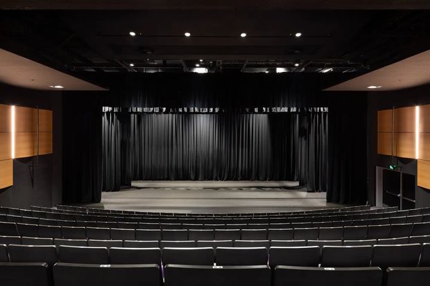 A dark, intimate theatre auditorium viewed from the rear, with rows of black upholstered seating facing a raised stage framed by heavy black curtains. Warm wood acoustic wall panels with vertical lighting accents line both sides, while recessed ceiling lights and visible stage rigging complete the formal performance space.
