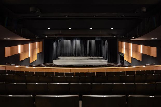 Interior view of a modern auditorium or theatre, looking toward a curtained stage from rows of dark upholstered seating. The space features black ceilings and walls, warm timber-toned acoustic wall panels, recessed spotlights, and a broad, gently curved balcony or seating arrangement.