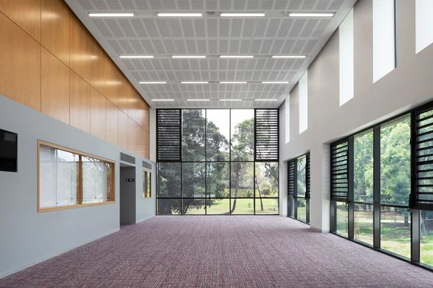 A spacious contemporary hall or multipurpose interior with a high gridded acoustic ceiling, extensive floor-to-ceiling glazing, black louvred window screens, pale grey walls, warm timber wall panelling, and mauve patterned carpet. The room opens visually to landscaped grounds through large windows and glazed doors, with strong natural light and a clean institutional or commercial character.