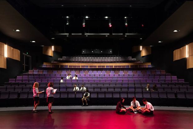 A dark, tiered theatre auditorium viewed from the stage, with rows of purple upholstered seating, a curved rear balcony, black ceiling and walls, warm wood acoustic wall panels, and theatrical stage lighting casting red illumination across the glossy stage floor. Several people are scattered among the seats and on the stage.
