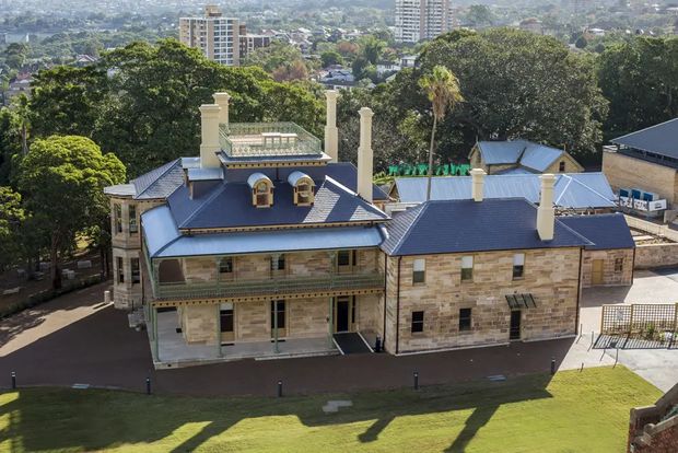 Aerial view of a substantial historic stone building with multiple blue-grey pitched roofs, tall chimneys, dormer windows, verandahs, and decorative balustrades. The building sits within landscaped grounds on a sloping urban site, with a dense city neighbourhood and high-rise buildings visible in the background.