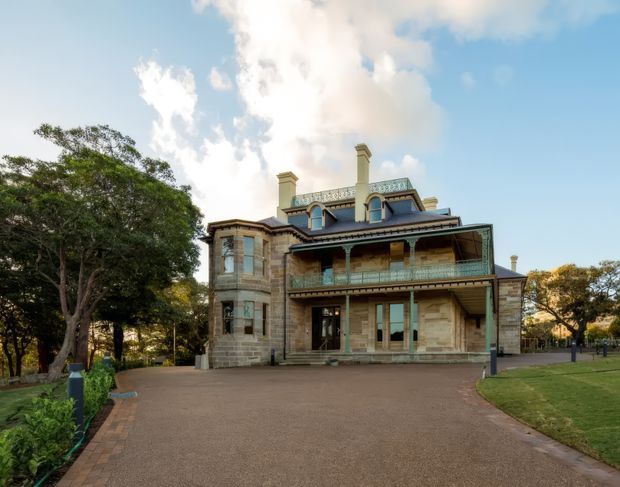 Exterior view of a substantial historic masonry residence set within landscaped grounds. The two-storey building features sandstone or rendered stone walls, ornate verandahs and balustrades, tall chimneys, decorative timber detailing, and a dark pitched roof. A broad driveway leads toward the front entrance, with mature trees and lawn surrounding the property.