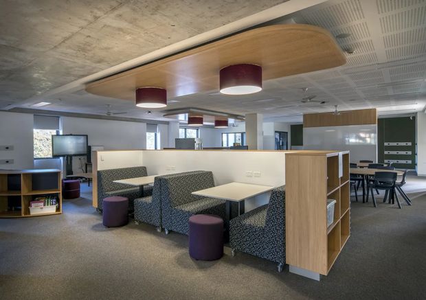 Open-plan contemporary commercial interior, likely a workplace or shared office lounge, with clustered upholstered booth seating, small tables, ottomans, timber room dividers and shelving, and a dining or collaboration area in the background. The space has exposed concrete ceilings, acoustic ceiling panels, integrated timber ceiling feature panels with pendant lights, carpeted flooring, and a neutral palette with muted purple accents.