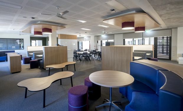 A bright contemporary commercial breakout and collaboration area with blue curved upholstered booth seating, round and organically shaped timber-look tables, purple ottomans, timber privacy partitions, and clustered café-style tables and chairs in the background. The space has grey carpet, a perforated acoustic ceiling, recessed lighting, sculptural pendant fixtures, and extensive interior glazing.