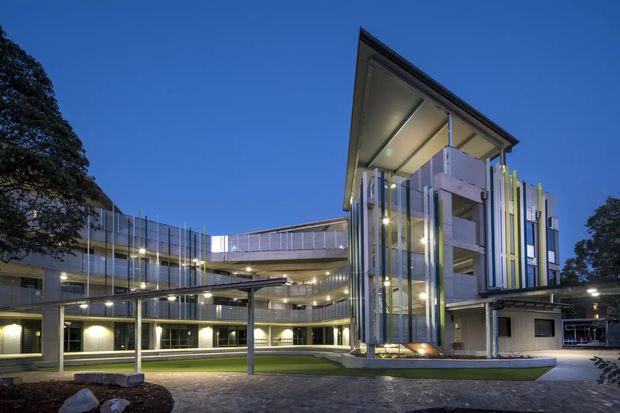 A contemporary multi-storey institutional or commercial building photographed at dusk. The complex features a prominent angular projecting roof, extensive glazing, layered horizontal floor plates, an illuminated courtyard, and a dramatic vertical facade screen composed of slender multicoloured fins. A covered walkway or canopy connects the building volumes, with landscaped paving and lawn in the foreground.