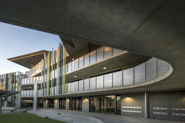 Exterior view of a contemporary multi-storey building with sweeping curved concrete canopies, exposed concrete soffits, extensive glazing, vertical multicoloured fins, and ground-level entry and garage-style doors. The landscaped foreground and surrounding sky are secondary to the architectural facade.
